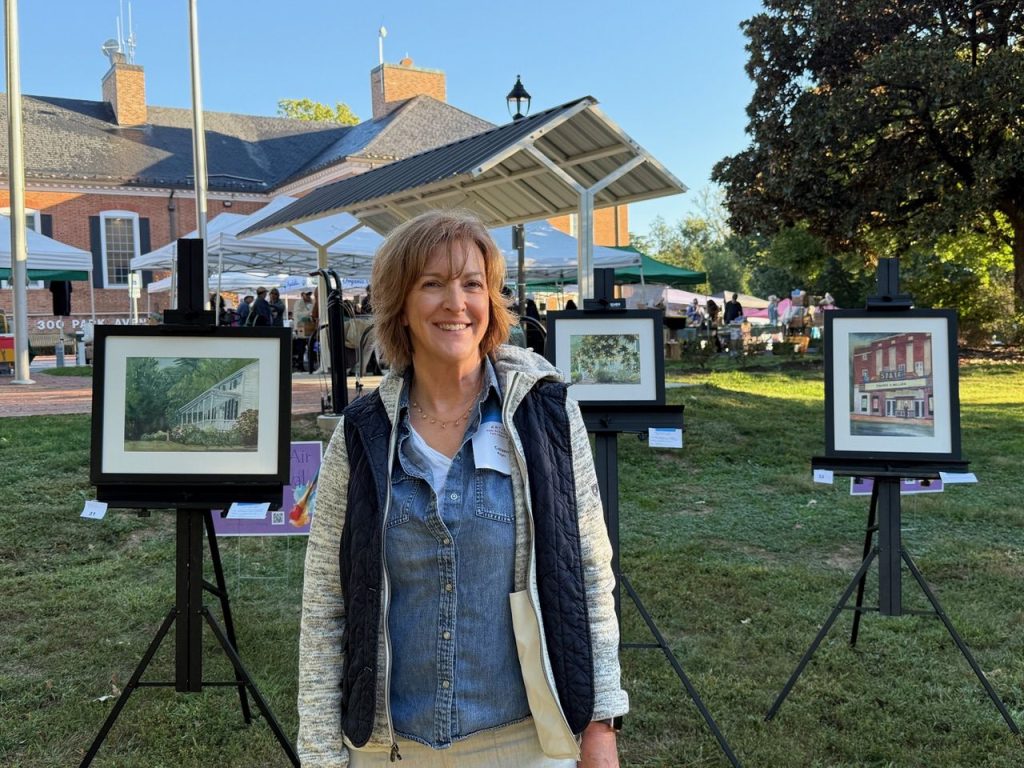 Image of artist Colleen Regan standing in the foreground with three of her paintings on easels behind her. Location is an outdoor art show.